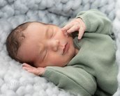 Neugeborenes Baby mit braunen Haaren, das in einem grünen Strickpullover auf einem weichen, grauen Hintergrund schläft.