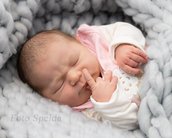 Neugeborenes Baby mit geschlossenen Augen, das in einer grauen Decke liegt und mit einem Finger seine Nase berührt.