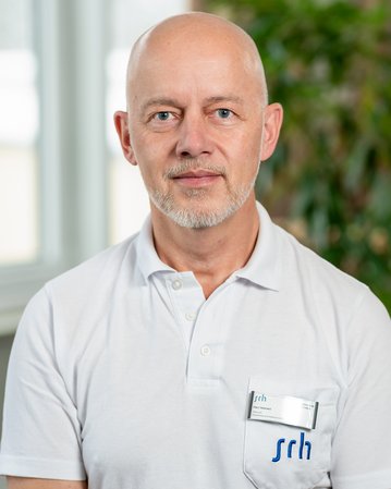 Ein Mann mit Glatze und Bart trägt ein weißes Polohemd. Er schaut direkt in die Kamera, während er vor einer Pflanze steht. Ein Namensschild ist sichtbar.