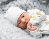 Neugeborenes Baby mit einem weißen Mützchen und einem Schlafanzug mit Winnie-Pooh-Muster, das auf einer grauen, strukturierten Decke liegt. Das Baby schaut direkt in die Kamera.