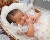 Neugeborenes Baby schläft in einem Korb, trägt ein zartes, blütenverziertes Kleid und hat die Finger im Mund.