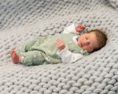 Ein schlafendes Baby in einem grünen Overall liegt auf einer grauen, gestrickten Decke.