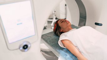Eine Patientin liegt auf einer Liege in einem CT-Scanner. Der Scanner hat eine runde Öffnung und ist Teil einer medizinischen Einrichtung. Ein Bildschirm zeigt Informationen an.