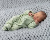 Ein schlafendes Neugeborenes in einem grünen Overall mit Punkten liegt auf einer grauen, gestrickten Decke. Das Baby hat die Hände in der Nähe des Gesichts und wirkt friedlich.