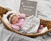 Neugeborenes Mädchen in einem Korb, trägt ein rosa Oberteil und einen blumigen Rock, mit einer Blütenkrone. Ein Schild sagt "Willkommen Martha".