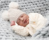 Ein schlafendes Neugeborenes in einem weißen Strickpullover und einer Mütze mit Pompons, eingekuschelt in eine graue Decke.
