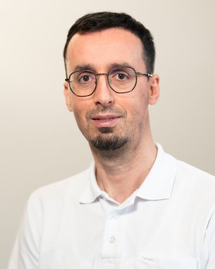 Ein Mann mit Brille und kurzem, dunklem Haar trägt ein weißes Polohemd mit einem blauen Logo auf der Brust. Er steht vor einem neutralen Hintergrund und schaut direkt in die Kamera.