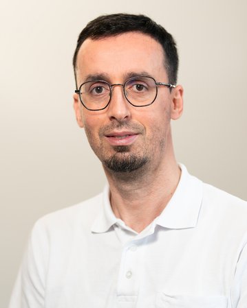 Ein Mann mit Brille und kurzem, dunklem Haar trägt ein weißes Polohemd mit einem blauen Logo auf der Brust. Er steht vor einem neutralen Hintergrund und schaut direkt in die Kamera.