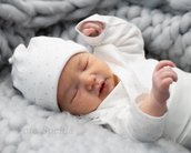 Ein schlafendes Baby in einem weißen, gepunkteten Outfit und einer Mütze liegt auf einer grauen, strukturierten Decke. Das Baby hat die Augen geschlossen und die Hände leicht angehoben.