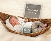 Neugeborenes Baby in einem geflochtenen Korb, trägt ein weißes Oberteil und gemusterte Hosen. Ein Schild mit "Willkommen Ben" und einem Datum steht daneben.