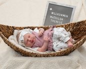 Neugeborenes Baby in einer Korbwiege, trägt ein rosa Oberteil und eine Blumenhose, neben einem Schild mit dem Namen "Mathilda Therese".