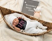 Neugeborenes Baby in einem geflochtenen Korb, trägt einen schwarzen Body mit dem Schriftzug "Mini" und eine weiße Mütze. Neben dem Baby steht ein Schild mit den Worten "Willkommen Mailey Joanna" und ein Stofftier.