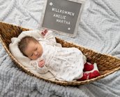 Neugeborenes Baby in einem geflochtenen Korb, trägt ein weißes Kleid und rote Socken. Ein Schild mit dem Text "Willkommen Amelie Martha" steht daneben.