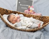 Neugeborenes Baby liegt in einem Korb, umgeben von einer Decke und einem Spielzeug, mit einem Schild, das "Willkommen Frieda" zeigt.