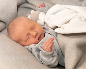 Ein schlafendes Baby in einem grauen Strickpullover, eingekuschelt in eine Decke, hält ein Kuscheltier in der Hand.