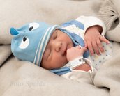 Ein schlafendes Baby trägt eine blaue Mütze mit Augen und hält seinen Daumen. Es liegt in einer weichen Decke.