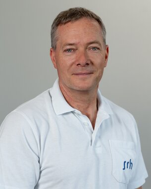 Mann mit kurzen Haaren trägt ein weißes Poloshirt mit einer Brusttasche. Der Hintergrund ist neutral und hell.