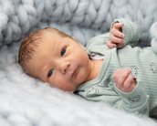 Neugeborenes Baby mit kurzen, hellbraunen Haaren und blauen Augen, das in einem grauen, gestrickten Überwurf liegt. Es trägt einen hellgrünen, gestreiften Overall mit Tiermotiven.