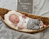 Neugeborenes Baby liegt in einem geflochtenen Korb, trägt ein T-Shirt mit dem Text „Mein erster Weihnachten“ und gestreifte Hosen. Ein Schild mit „Willkommen Jaro Josef“ steht daneben.