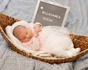 Neugeborenes Baby in einem Korb, trägt ein rosa Outfit und liegt auf einer Decke. Ein Schild mit "Willkommen Alicja" steht daneben.