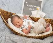 Neugeborenes Baby liegt in einem Korb, trägt ein T-Shirt mit Tiermotiven und schaut zur Seite. Ein Schild mit "Willkommen Enno" ist im Hintergrund.