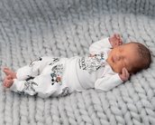 Ein schlafendes Neugeborenes liegt auf einer grauen, gestrickten Decke. Es trägt einen weißen Schlafanzug mit Mickey-Maus-Motiven. Die Hände sind entspannt und die Augen sind geschlossen.