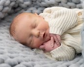 Ein schlafendes Neugeborenes in einem weißen Strickpullover, eingekuschelt in eine graue, weiche Decke.