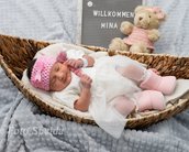Neugeborenes Mädchen in einem Korb, trägt ein weißes Kleid und eine rosa Mütze. Neben ihr steht ein Plakat mit "Willkommen Mina" und einem Plüschbären.