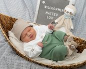 Neugeborenes Baby in einem Korb, trägt einen Hut und grüne Hose. Neben ihm steht ein Schild mit "Willkommen Matteo Loui" und ein Plüschtier.