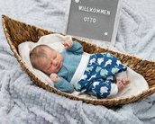 Ein schlafendes Baby liegt in einem geflochtenen Korb, eingekuschelt in eine weiße Decke. Es trägt einen blauen Oberteil und eine Hose mit einem Muster aus Elefanten. Ein Schild im Hintergrund zeigt den Text "Willkommen Otto".
