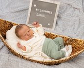 Neugeborenes Baby liegt in einem Korb, trägt ein weißes Oberteil und grüne Hosen. Ein Schild mit der Aufschrift "Willkommen Daniel" steht daneben.