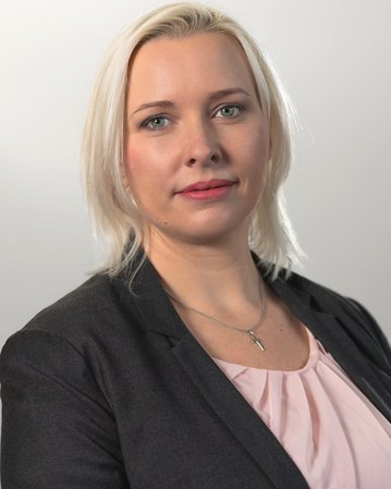 Eine Frau mit blonden, schulterlangen Haaren trägt einen schwarzen Blazer und ein hellrosa Oberteil. Sie schaut direkt in die Kamera, mit einem neutralen Gesichtsausdruck und einem dezenten Schmuckstück um den Hals.