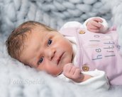 Neugeborenes Baby mit braunen Haaren in einem rosa Overall, das auf einer grauen Decke liegt und in die Kamera schaut.