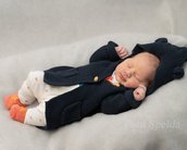 Ein schlafendes Neugeborenes in einem dunkelblauen Strickmantel mit Kapuze, weißen Strumpfhosen und orangefarbenen Socken auf einem grauen Hintergrund.