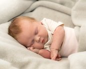 Ein schlafendes Baby mit blonden Haaren, eingekuschelt in eine graue Decke, liegt auf der Seite und hat die Hände unter dem Kopf.