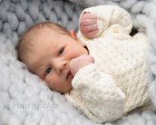 Ein neugeborenes Baby mit blauen Augen liegt in einer weichen, grauen Decke und trägt einen weißen, gestrickten Pullover.