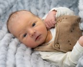 Neugeborenes Baby in einem weichen, grauen Strickmaterial, trägt einen braunen Overall mit Knöpfen und schaut direkt in die Kamera.