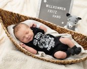 Neugeborenes Baby schläft in einem Korb, trägt einen schwarzen Body mit einem Aufdruck. Neben dem Baby liegt ein graues Stofftier und ein Schild mit dem Text "Willkommen Fritz Hans".