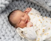 Ein schlafendes Baby mit einem sanften Lächeln, gekleidet in einem weißen Outfit mit Blumenmuster, liegt auf einer grauen, gestrickten Decke.