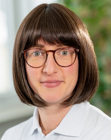 Frau mit braunem Bob-Haar und Brille trägt ein weißes Polohemd mit dem Logo "srh". Sie steht in einem hellen Raum mit Pflanzen im Hintergrund.