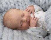 Ein schlafendes Neugeborenes mit blonden Haaren, eingekuschelt in eine graue Decke, trägt einen weißen Strampler mit kleinen Blumen.
