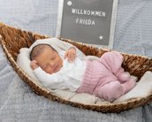 Neugeborenes Baby in einem Korb, eingekuschelt in eine Decke, trägt einen weißen Oberteil und rosa Hosen. Ein Schild mit "Willkommen Frieda" steht daneben.