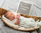 Neugeborenes Baby in einer Korbwiege, umgeben von Decken, mit einem Schild, das "Willkommen Gabriel" zeigt.
