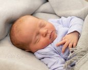 Ein schlafendes Baby in einem hellblauen Oberteil, eingewickelt in eine graue Decke, mit einer entspannten Körperhaltung.