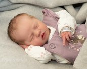 Ein schlafendes Baby in einem lila Overall mit Blumenmuster, auf einer grauen Decke liegend, mit einer Hand unter dem Kopf.