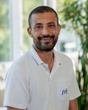 Ein Mann mit kurzem Bart und lächelndem Gesicht trägt ein weißes Polohemd. Er sitzt in einem hellen Raum mit Pflanzen im Hintergrund.