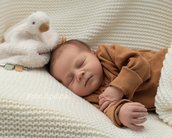 Ein schlafendes Baby in einem braunen Outfit liegt auf einer weißen Decke, neben einem plüschigen Spielzeug.