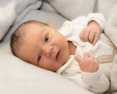 Neugeborenes Baby liegt auf einer Decke, trägt einen weißen Overall und schaut direkt in die Kamera.