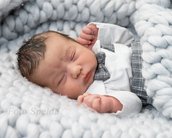 Ein schlafendes Neugeborenes in einem weißen Hemd mit einer grauen Fliege, eingewickelt in eine weiche, graue Decke. Das Baby hat dunkles, lockiges Haar und liegt auf einer strukturierten, grauen Unterlage.