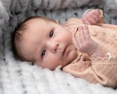 Ein Neugeborenes mit braunen Augen und leichtem Lächeln, gekleidet in einen hellen Pullover, liegt auf einer weichen Decke.
